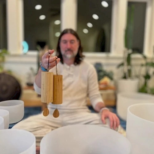 A bearded man in white robes sits cross-legged indoors, holding a wind chime or bamboo chime, surrounded by white bowls or bowls and plants.