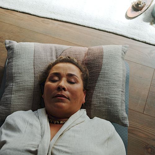 A woman peacefully lying on a pillow with eyes closed, wearing a white robe, relaxing indoors on a wooden floor with a cozy setup nearby.