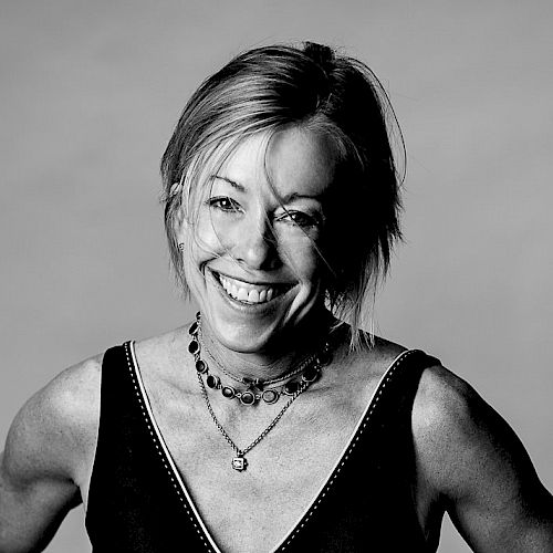 A smiling woman with short hair, wearing a sleeveless top and layered necklaces, posing confidently.