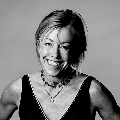 A smiling woman with short hair in a sleeveless top, necklace, and a confident pose.