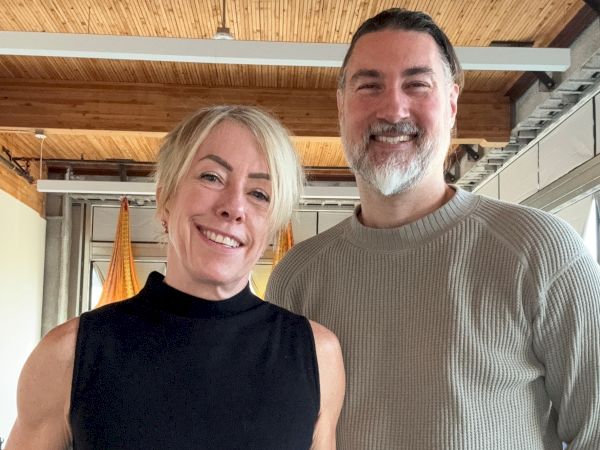 A smiling couple stands together indoors, woman in a sleeveless black top and man in a light gray sweater, both casually dressed and posing.