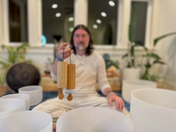 A man sits cross-legged indoors, holding a small wooden wind chime or percussion instrument, with hanging bars and tiny bells. End.