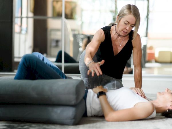 A woman in a black sleeveless topProviding chest compressions? No, she&rsquo;s pressing on a man&rsquo;s chest during a first-aid or CPR training session on a gym floor, with cushions nearby. ends with period