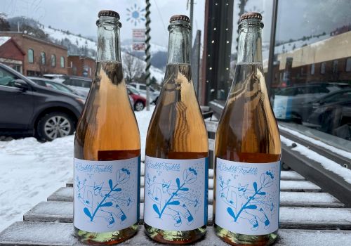 Three small glass bottles with light pink to amber liquid, labeled with blue botanical designs, on a snowy outdoor bench, reflective storefronts nearby.