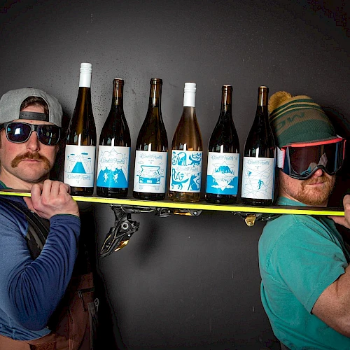 Two men wear silly hats and sunglasses, balancing a skateboard loaded with several wine bottles between them, posing for a quirky shot.