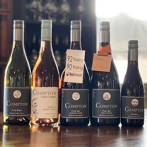 A row of five wine bottles on a wooden surface, labels read Compton and other vintages, with tags hanging from two bottles, bright light in the background.