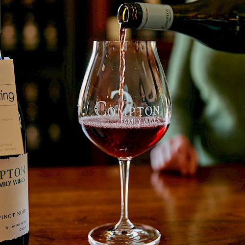 A wine glass being poured with red wine from a bottle, on a wooden bar.