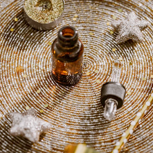 An amber dropper bottle, a small bulb syringe, and tiny ornaments on a glittery, spiraled surface with stars and festive decor.