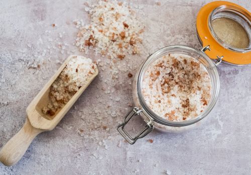 A jar of crumbly salt or sugar scrub with a scoop and scattered grains, on a textured surface, ready for use.