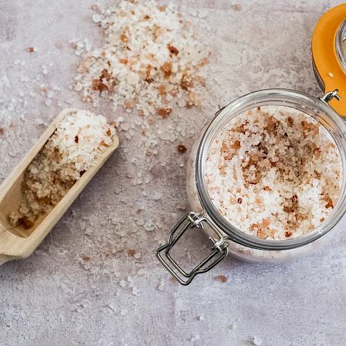 A jar of crumbly salt or sugar scrub with a scoop and scattered grains, on a textured surface, ready for use.