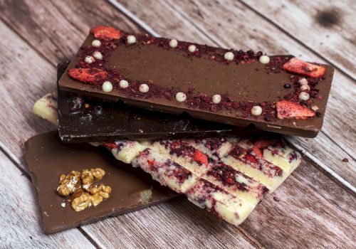 Assorted chocolate bars with toppings (berries, white bits, nuts) stacked on a wooden surface, including a white chocolate variety with berry swirls.