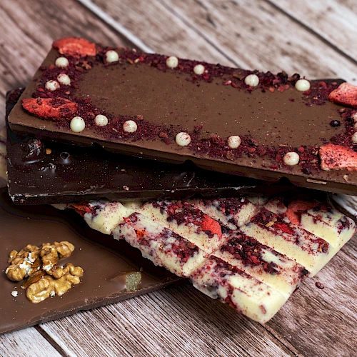 Assorted chocolate bars with toppings (berries, white bits, nuts) stacked on a wooden surface, including a white chocolate variety with berry swirls.