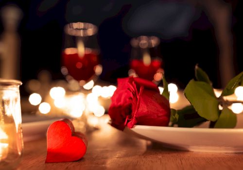 A romantic table setting with a red rose on a plate, a small red heart, candles, and dim bokeh lights in the background, creating a cozy vibe.