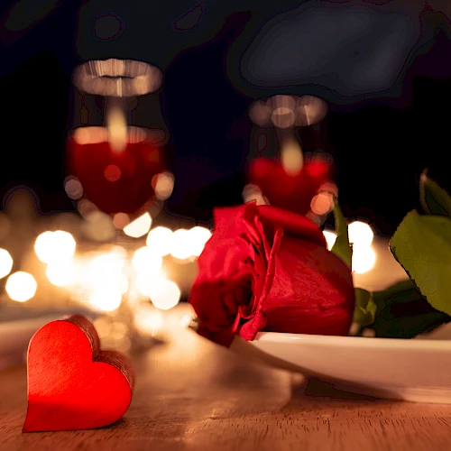 A romantic table setting with a red rose on a plate, a small red heart, candles, and dim bokeh lights in the background, creating a cozy vibe.