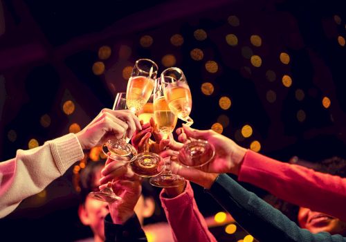 People clinking champagne flutes in a festive toast at a party, with warm bokeh lights in the background.
