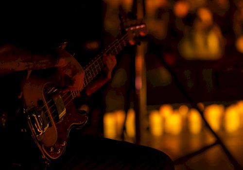 A dimly lit stage with a bass guitar resting on a musician&rsquo;s knee; warm, blurry background lights hint at an audience or venue.