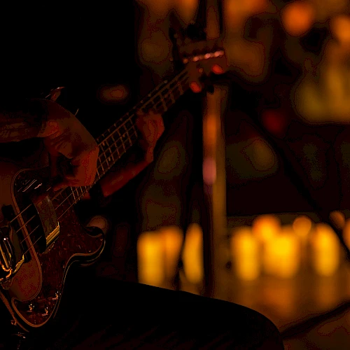 A dimly lit stage with a bass guitar resting on a musician’s knee; warm, blurry background lights hint at an audience or venue.
