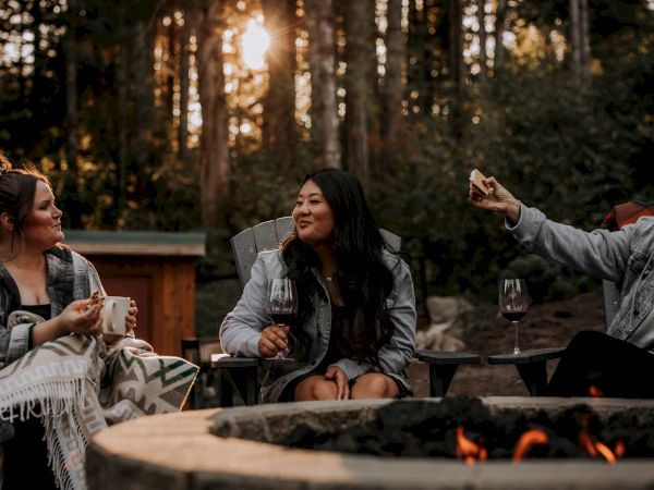 Three friends sit around a fire pit outdoors at dusk, chatting and sipping drinks, cozy blankets around them, trees in the background.