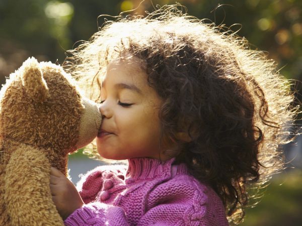 A young girl in a pink sweater shares a kiss with a brown teddy bear outdoors, sunlight and trees in the background.