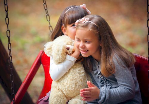 Two smiling girls hug a fluffy stuffed bear on a swing, sharing a warm moment outdoors.