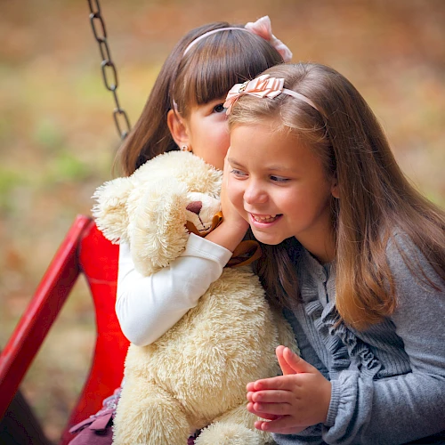 Two smiling girls hug a fluffy stuffed bear on a swing, sharing a warm moment outdoors.