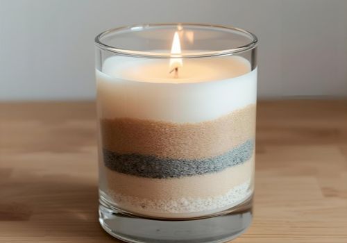 A lit, layered candle in a clear glass jar sits on a wooden surface, showing creamy white, tan, and dark brown sandy layers.