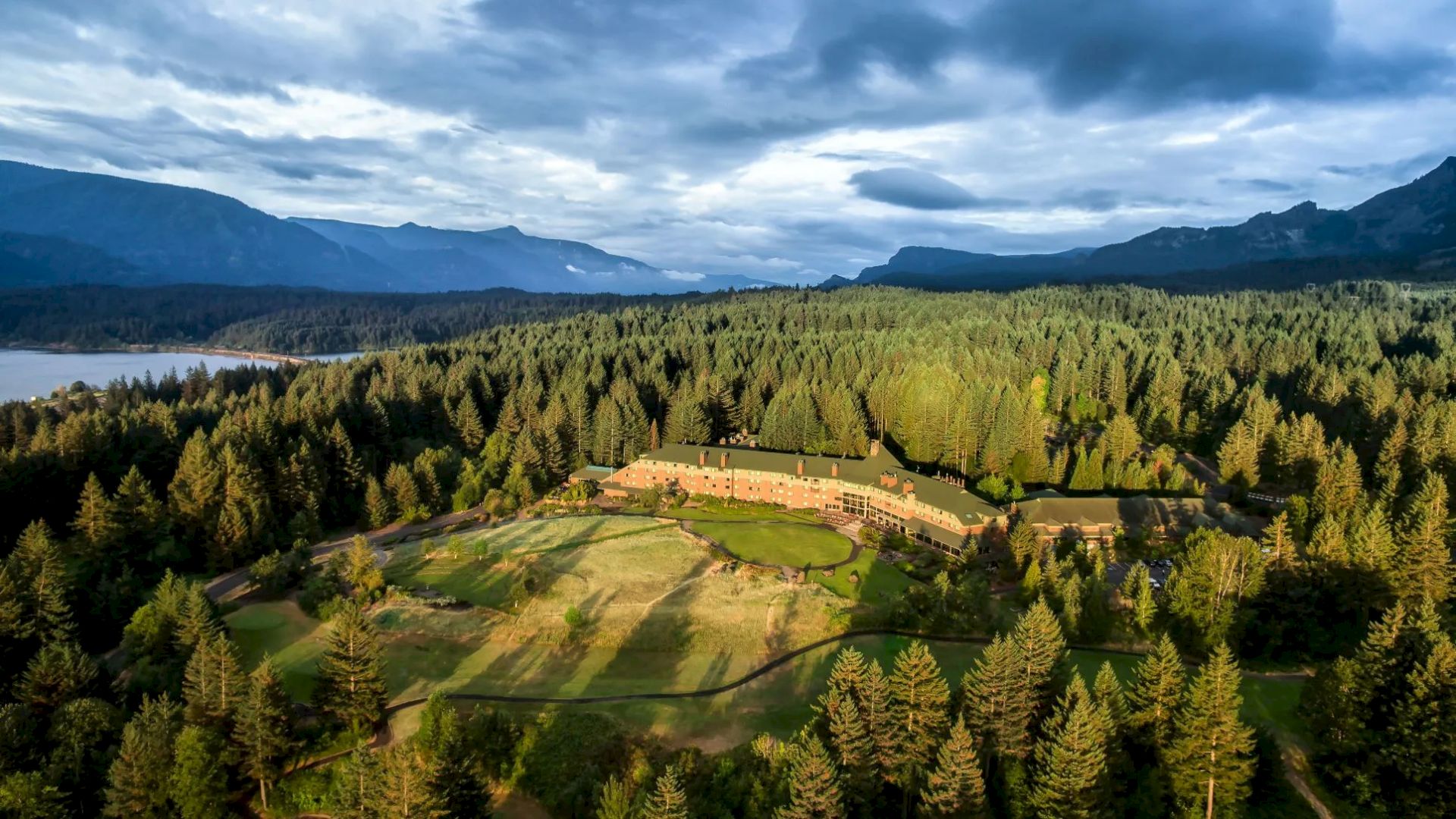 A large building is nestled in a forested area, surrounded by trees and mountains, with a view of water in the distance.