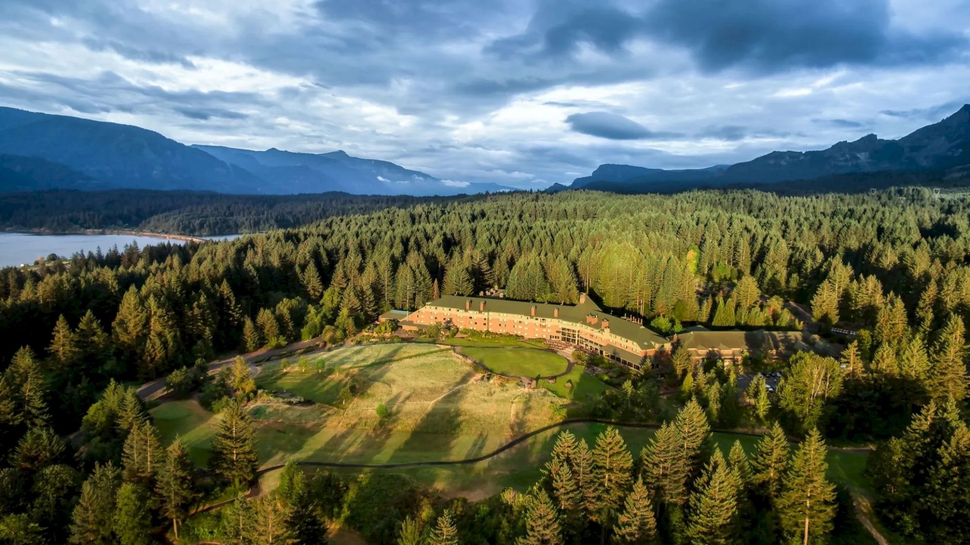 A large building is nestled in a forested area, surrounded by trees and mountains, with a view of water in the distance.