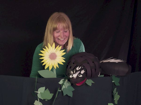 A smiling woman peers over a stage with a sunflower prop and a black cartoonish creature peeking from a leafy backdrop, sunny and playful.