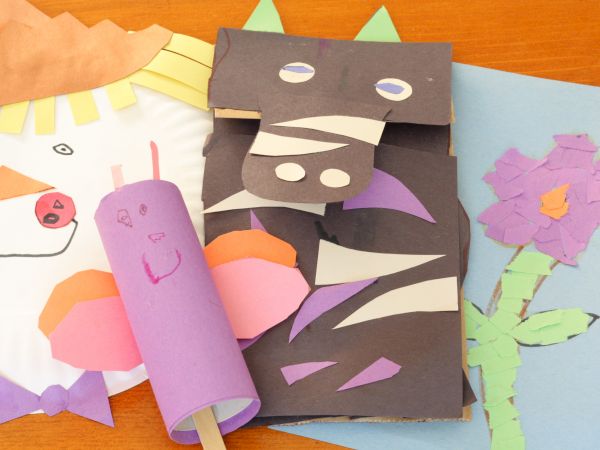 A colorful collage of paper crafts: a purple rolled tube with a face, a dark mask-like figure with cutouts, a purple flower + stem card, and a light blue card with a pink flower, all on a wooden surface.