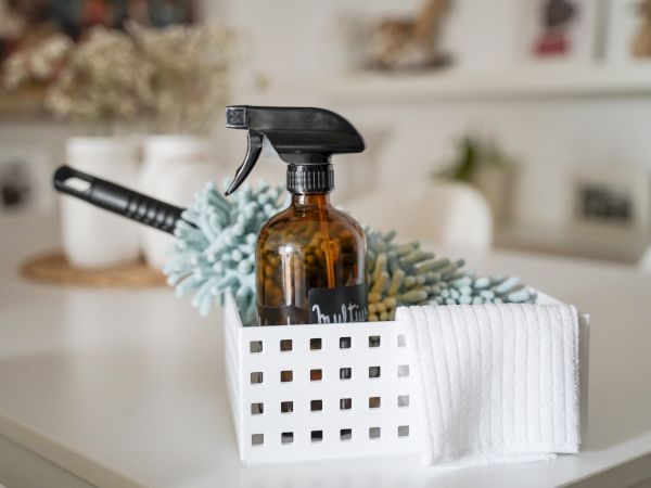 A white basket holds a brown spray bottle, a handheld vacuum, and a folded towel on a tidy bathroom counter.