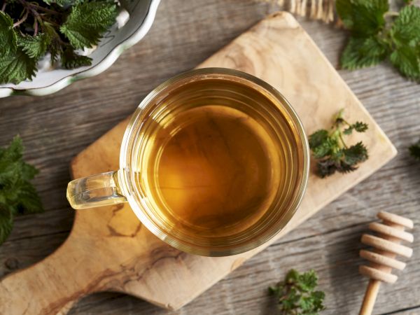 A cup of tea on a wooden board with herbs, ready to sip, surrounded by greenery and a honey dipper.