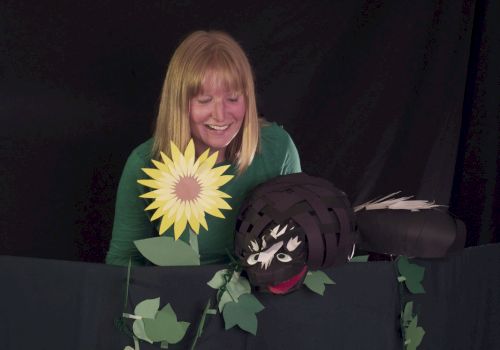 A smiling woman with a sunflower prop and a black puppet head peeking over a green ivy-covered backdrop, in a playful puppetry setup.