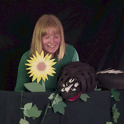 A smiling woman with a sunflower prop and a black puppet head peeking over a green ivy-covered backdrop, in a playful puppetry setup.