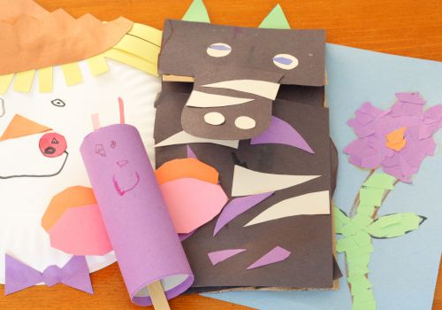 Colorful handmade crafts on a table: a purple paper roll creature, a dark folded dragon/monster with cutouts, a pink smiley face, and a blue card with a purple flower drawing, all surrounding a light-colored fan shape.