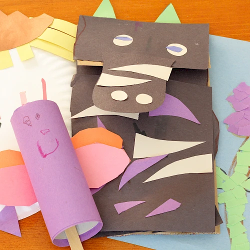 Colorful handmade crafts on a table: a purple paper roll creature, a dark folded dragon/monster with cutouts, a pink smiley face, and a blue card with a purple flower drawing, all surrounding a light-colored fan shape.