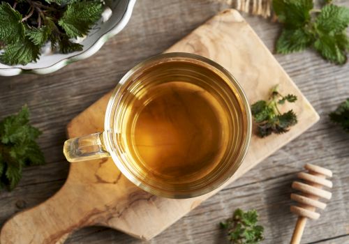 A cup of golden herbal tea sits on a wooden board with greens, a honey dipper, and fresh herbs around, cozy and rustic.