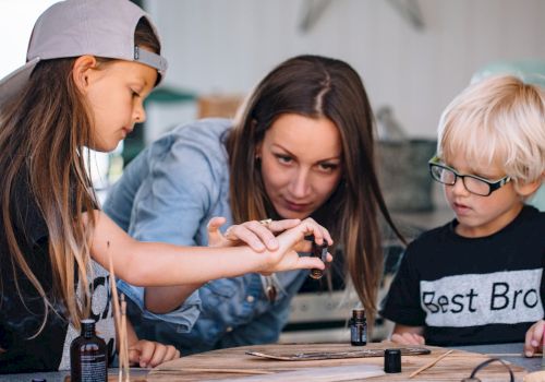 Three kids are painting at a table, with one girl guiding the younger pair as they focus on tiny details.