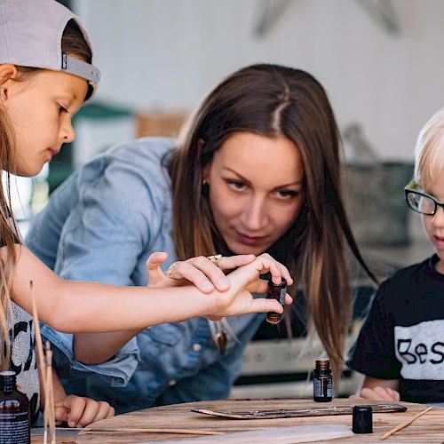Three kids are painting at a table, with one girl guiding the younger pair as they focus on tiny details.