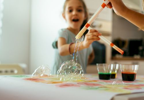 A child is playing with bubbling homemade slime or science experiment using droppers and colored liquids on a table, excitedly watching it spill.