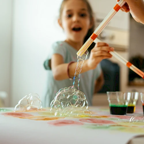 A child is playing with bubbling homemade slime or science experiment using droppers and colored liquids on a table, excitedly watching it spill.