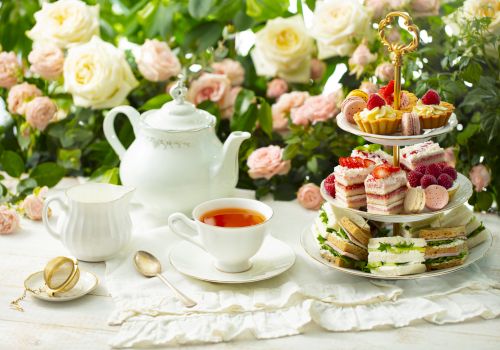 A delicate tea setup with a white teapot, cup, and saucer on a lace cloth, plus a three-tier pastry stand with assorted treats, against a floral backdrop. Top it at 140 characters, always ending the sentence.