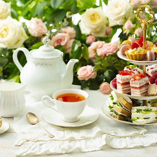 A delicate tea setup with a white teapot, cup, and saucer on a lace cloth, plus a three-tier pastry stand with assorted treats, against a floral backdrop. Top it at 140 characters, always ending the sentence.