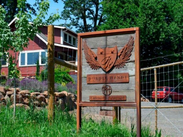 A wooden sign with an eagle emblem, set near a house and trees; a rocky yard and a gravel path in a sunny, rural setting.
