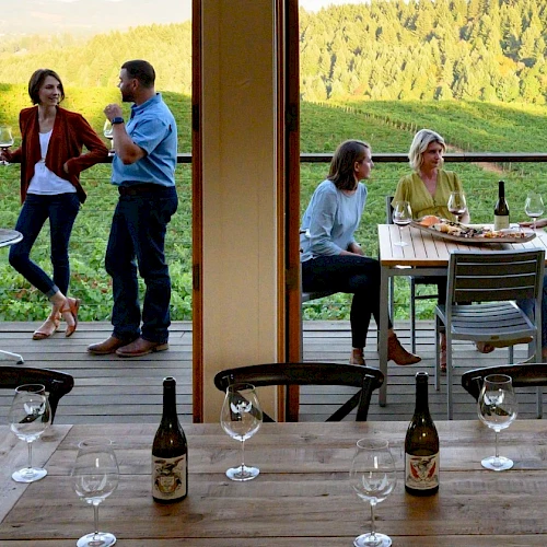 People enjoy a twilight winery vibe on a terrace: groups chat, toast, and sip wine as the sun sets over green hills behind them.