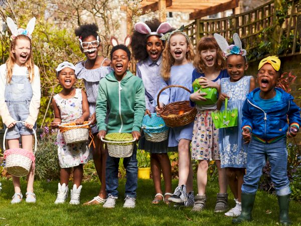 A group of kids wearing bunny ears and spring outfits pose in a garden, holding baskets and smiling for a festive photo.