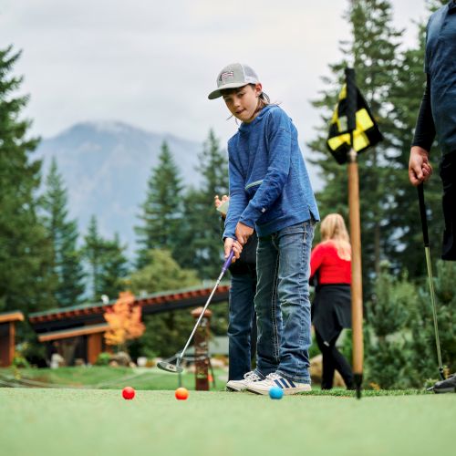 A person practices mini golf on a green outdoor course with a child aiming at a hole while others watch, set against trees and mountains.