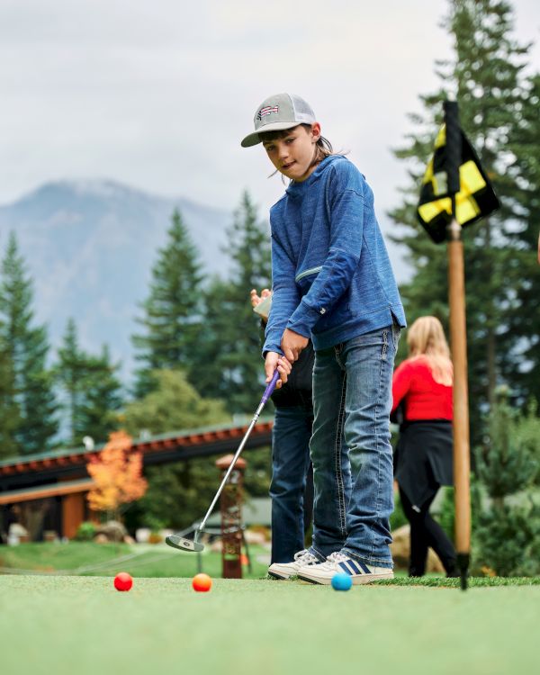 A person practices mini golf on a green outdoor course with a child aiming at a hole while others watch, set against trees and mountains.
