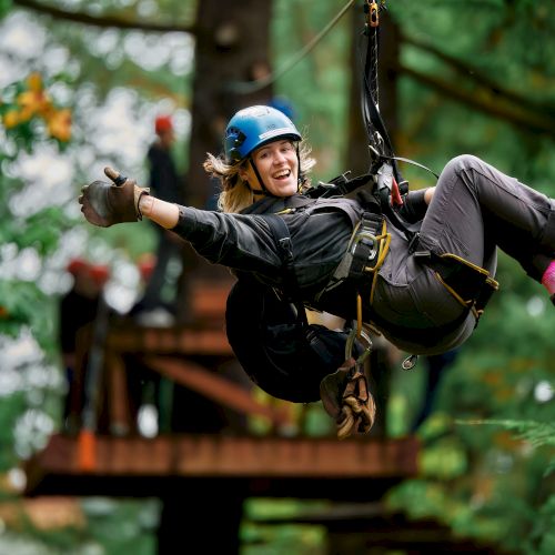 A person zip-lining through a forest, wearing a helmet and safety gear, smiling as they glide along a rope.