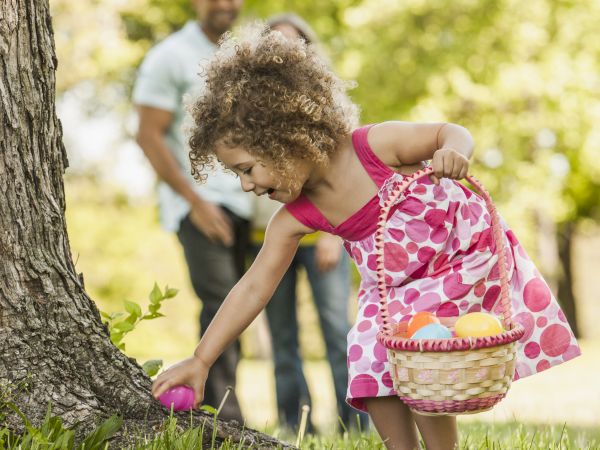 A young girl in a pink dress gathers something from the grass by a tree while a smiling man watches in the background; a sunny park scene.